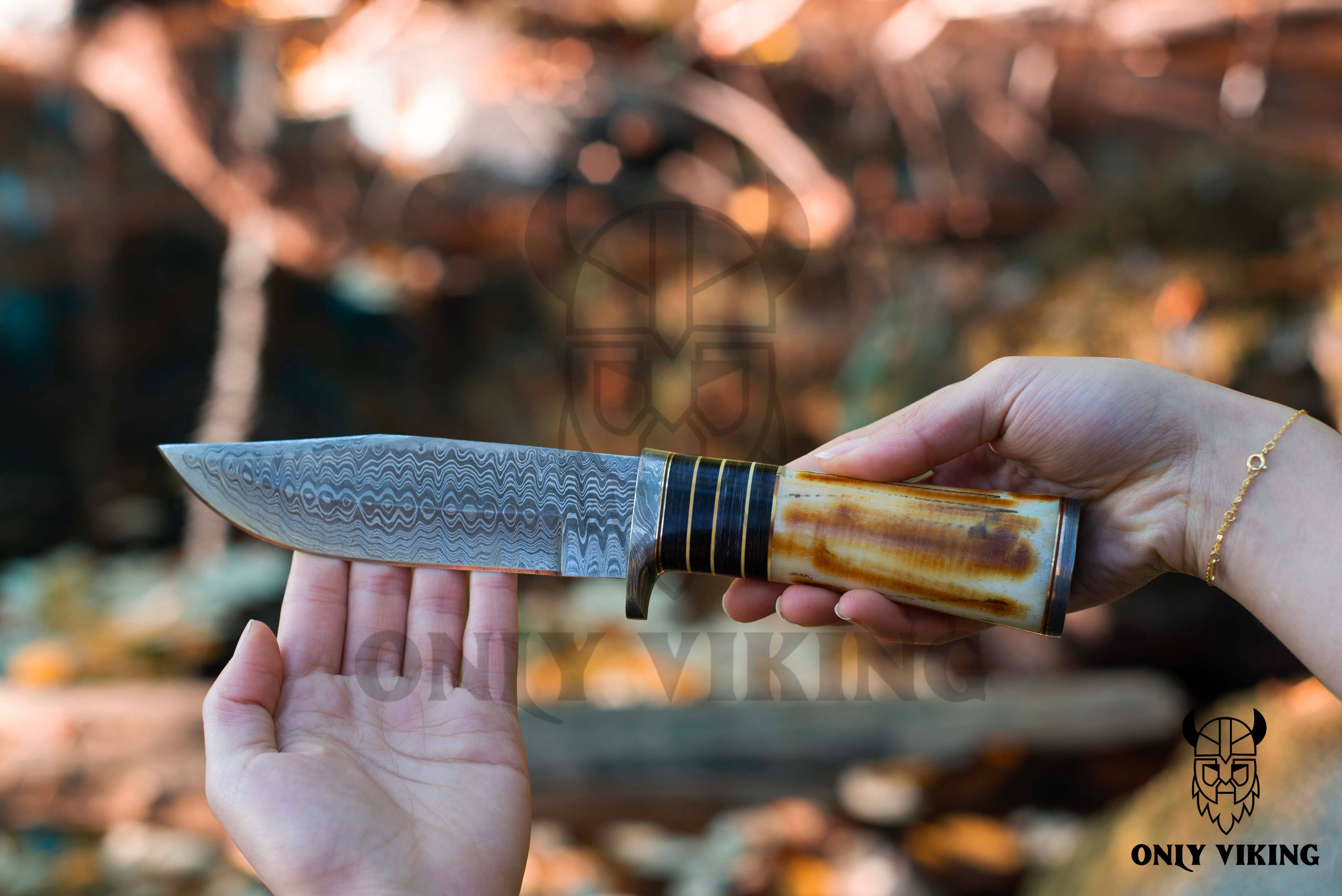 A person cautiously grips the Damascus Hunting Knife With Locking Leather Sheath, featuring a handcrafted patterned blade and ornate handle, perfect for outdoor activities. The knife gleams under sunlight, showcasing its intricate design.