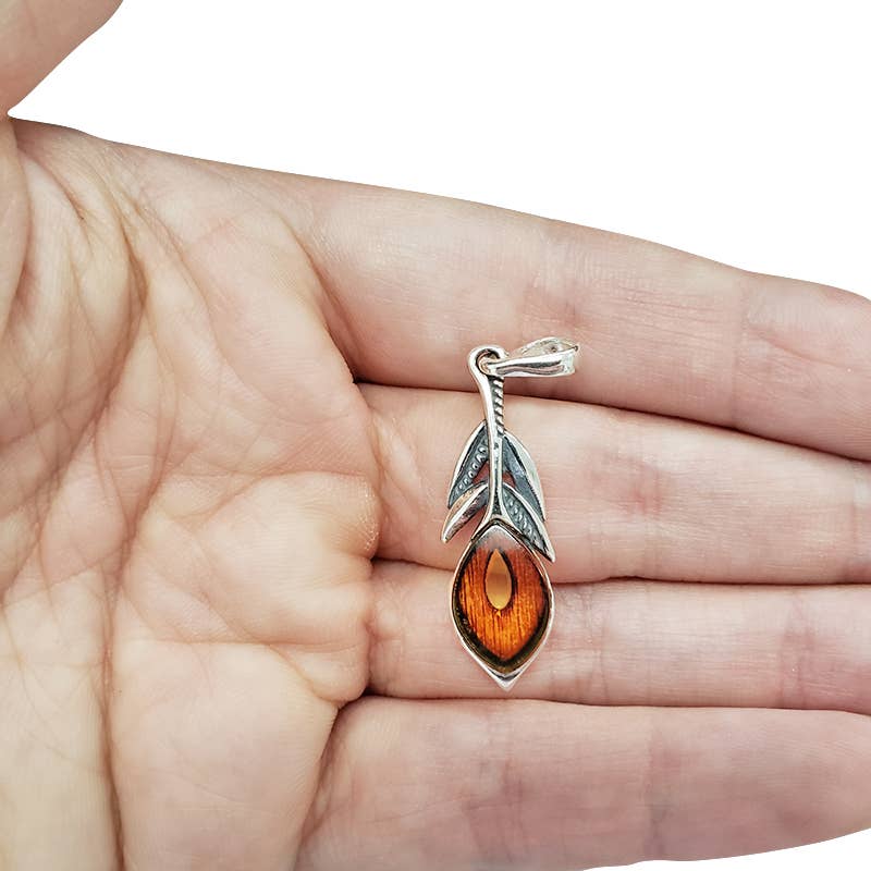 A hand holds a sterling silver pendant shaped like a peacock feather, featuring a genuine Baltic amber stone at its heart.