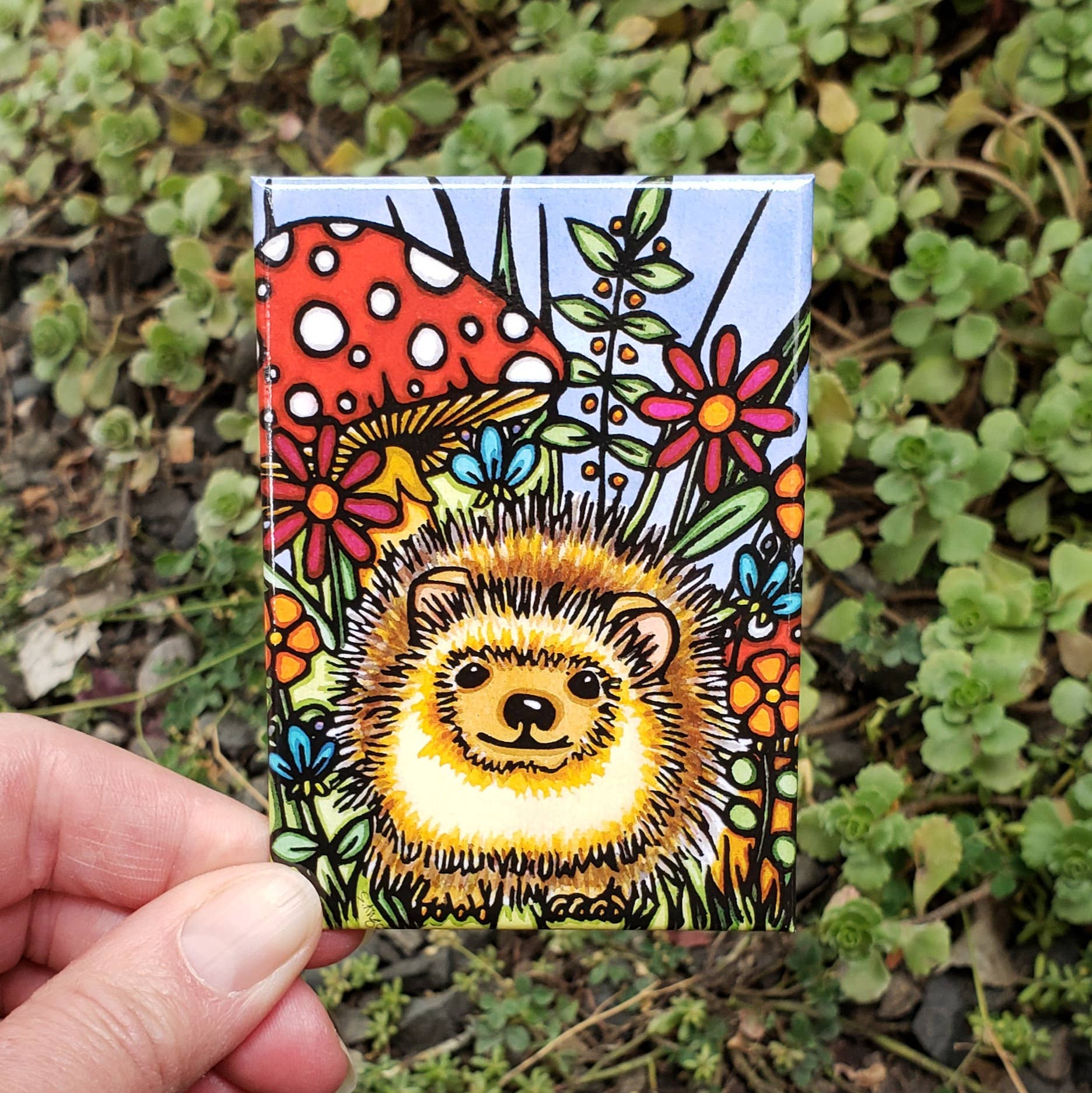 A hand holds the vibrant Hedgehog Magnet, featuring a detailed hedgehog amidst flowers, a mushroom, and lush greenery.