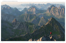 A person in a red jacket stands atop a mountain peak, surveying a vast range of jagged mountains under the cloudy Nordic sky, capturing the essence of *Wanderlust Nordics* and its legendary hiking routes.