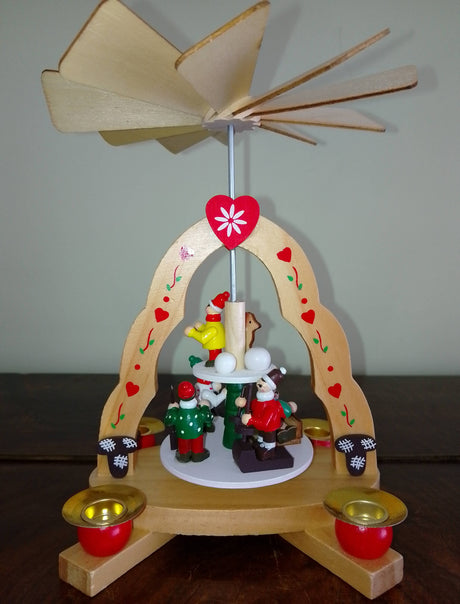 Handcrafted in Germany, the 7" x 6" Christmas Pyramid with "Kids" Candle Holder holds 4 candles and features festive figurines, heart motifs, and a fan rotor for a magical holiday display.