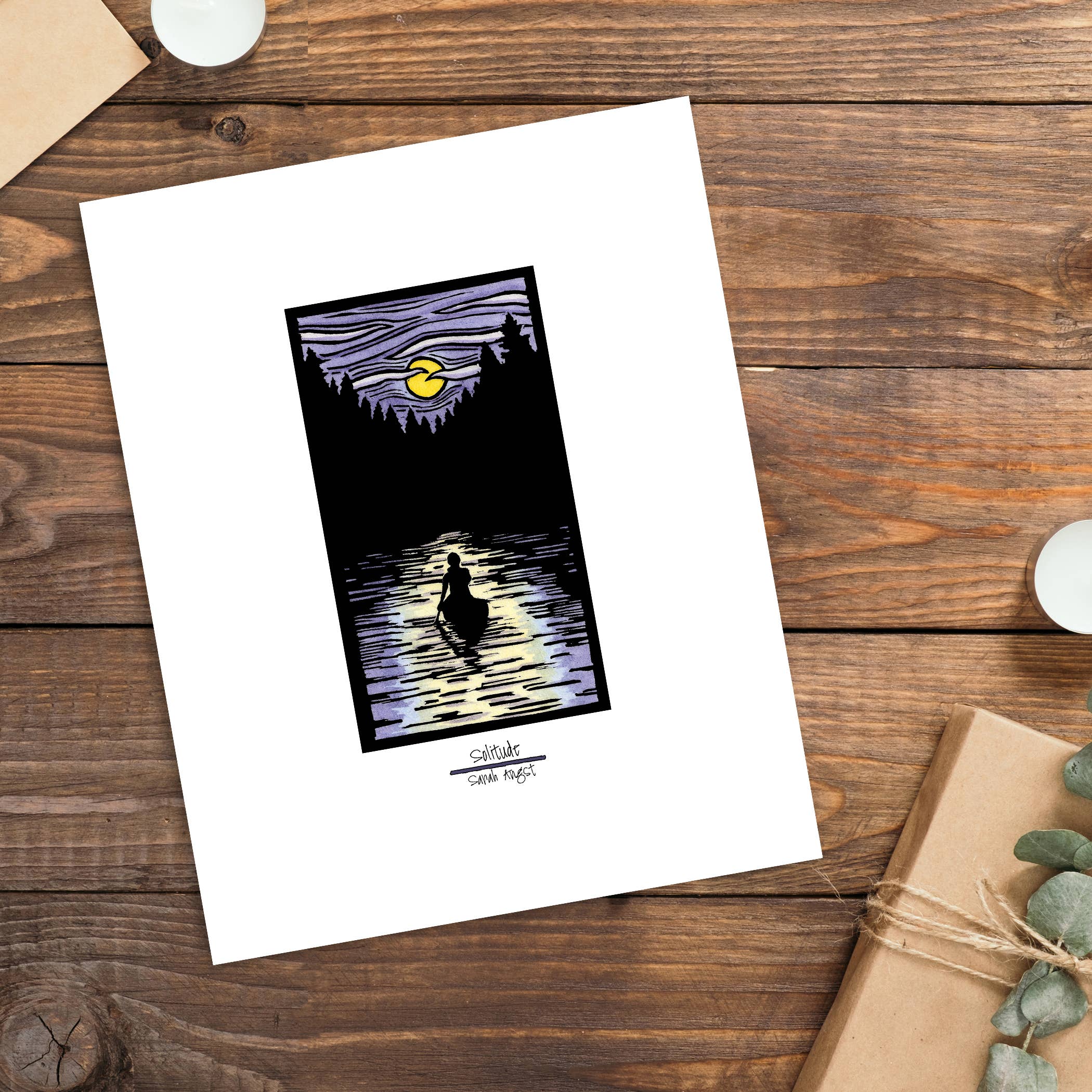 This 11"x14" framed Giclee print, "Solitude Canoe" by Sarah Angst, showcases a silhouetted figure paddling a canoe on a lake beneath a yellow moon, surrounded by trees. It is tastefully displayed on a wooden table alongside a gift box and candles.