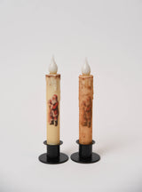 Two battery-operated LED taper candles, one cream and one textured brown, both with Santa Claus graphics in black metal holders.