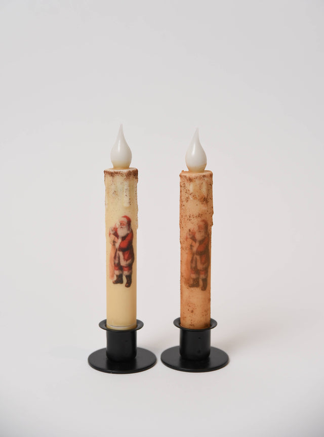 Two battery-operated LED taper candles, one cream and one textured brown, both with Santa Claus graphics in black metal holders.