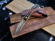 The Knife: Damascus Steel Pocket knife, featuring a Damascus steel blade and walnut wood handle, is displayed on wooden boards beside a brown leather sheath.