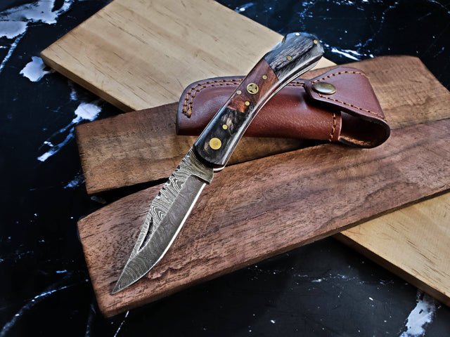 The Knife: Damascus Steel Pocket knife, featuring a Damascus steel blade and walnut wood handle, is displayed on wooden boards beside a brown leather sheath.