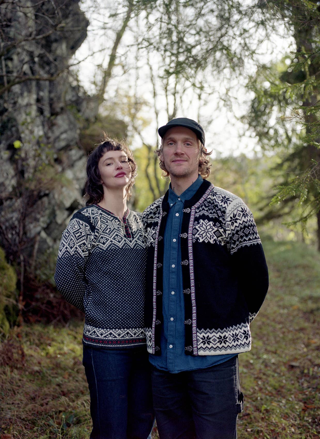 Two people stand outdoors in front of trees, both wearing traditional knitwear. One wears the Stavanger Norwegian Wool Cardigan in black, its classic pattern reflecting timeless Norwegian style. It appears to be daytime.