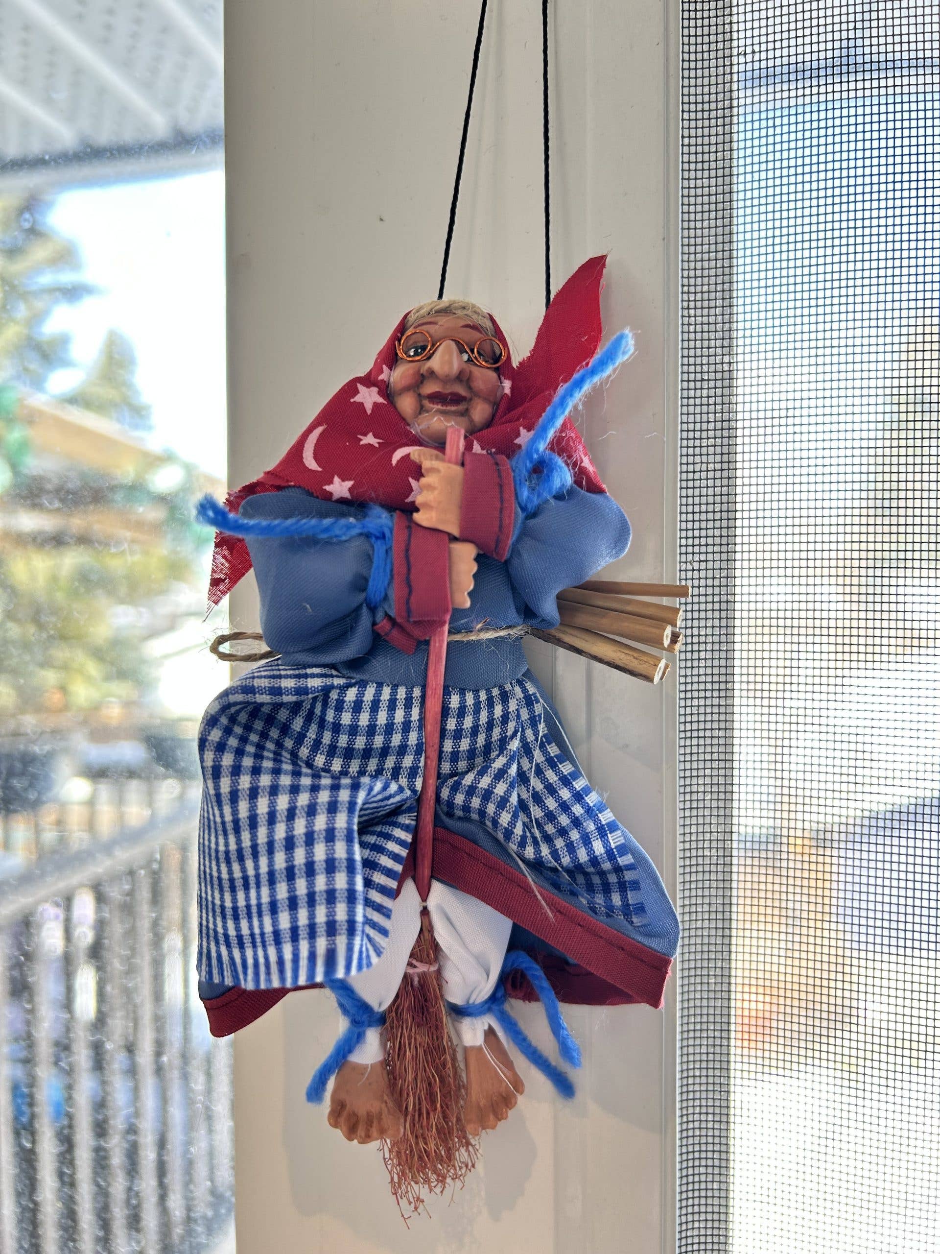 The Figurine: Opalina blue Kitchen Witch, 7" tall, wears blue clothes and a red headscarf, sitting on a small broomstick and hanging by a string near a window with snowy scenery in the background.