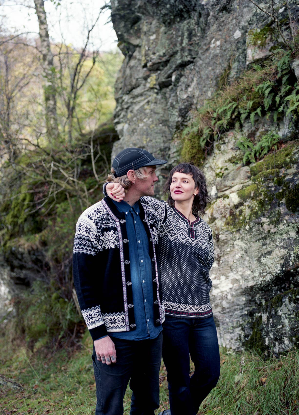 A man and woman wearing Stavanger Norwegian Wool Cardigan in black stand close together outdoors by a mossy rock wall, surrounded by greenery.