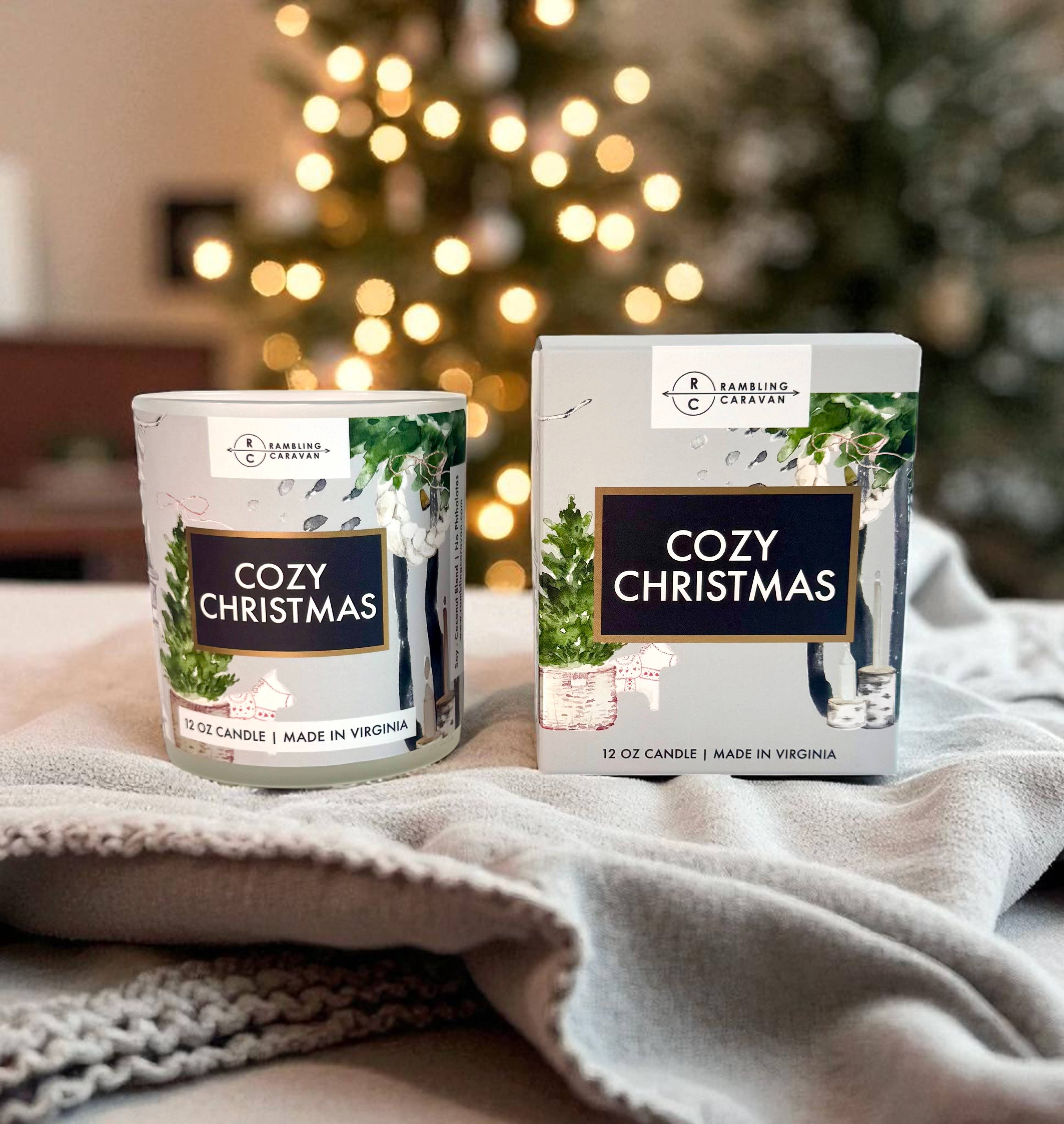 A lit and unlit 12 oz Cozy Christmas Candle with a matching box sit on a blanket, filling the air with inviting cinnamon vanilla scent, as a blurred Christmas tree softly glows in the background.