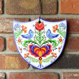 The Tile: Lovebirds Folk Art Cork Backed Ceramic Shield is a Nordic-designed wall decor featuring two blue birds, a heart, and flowers. This colorful ceramic plaque hangs beautifully on any wall.