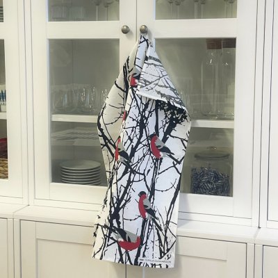 The Tea Towel: Bullfinches in Birch Trees "Domherrar" by Almedahls of Sweden, featuring black branches and red bird motifs, hangs from cabinet knobs before white glass-doored cupboards—a charming detail of Swedish kitchen decor.