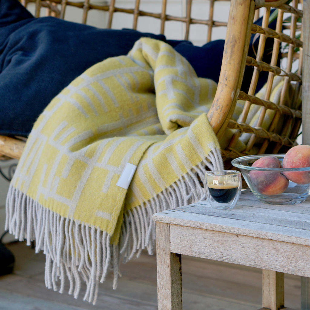 A wicker chair with the Pickepin Mustard blanket draped over it. A small table beside holds a double-walled glass with espresso and a bowl of peaches.