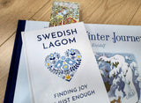 A book titled Swedish Lagom: Finding Joy in Just Enough rests atop another book and wood surface, with a postcard and the Bookmark: Carl Larsson 6-Piece Bookmark Collection (8.5" × 2.5" each) partially visible in the background.