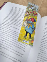An open book rests on a wooden surface with the Carl Larsson “The Apple Harvest” bookmark (8.5" × 2.5") placed between its pages, adding an elegant touch of Scandinavian artistry.