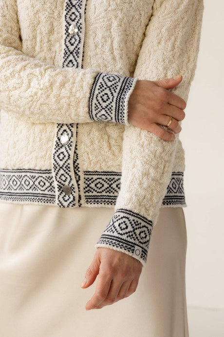 A person wearing the Bryggen Women's Norwegian Cardigan in white, featuring black geometric patterns on the cuffs and button band, paired with a cream skirt.
