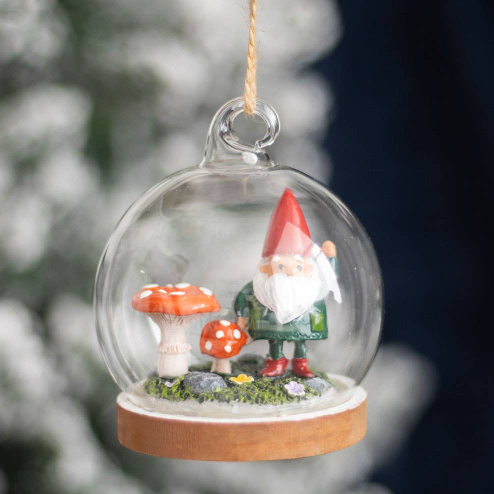 Introducing the "Ornament: Gnome Glass Dome," a charming decoration showcasing a small gnome and two mushrooms, all beautifully encased in a delicate glass dome suspended by a string.