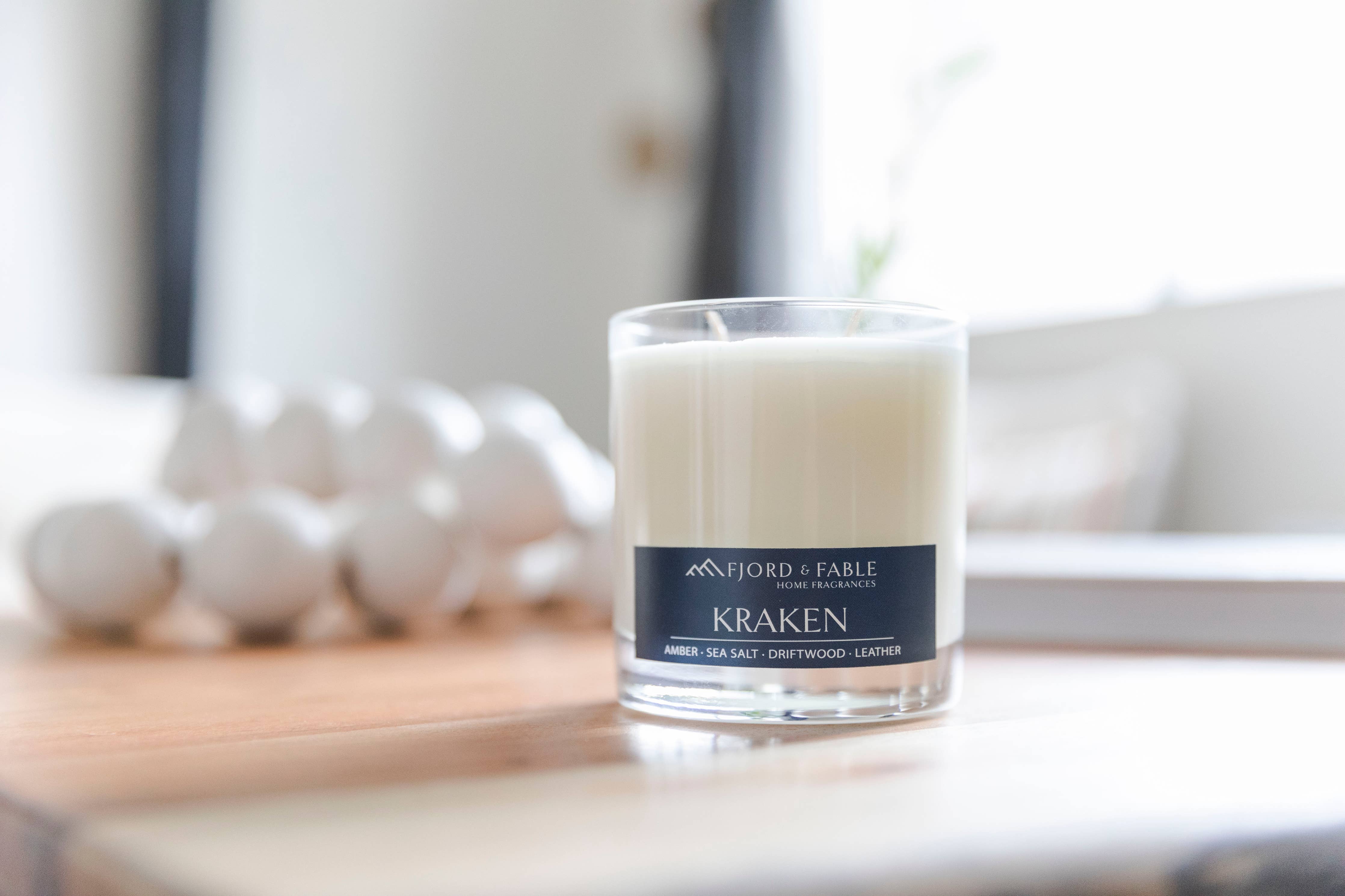 A white Fjord & Fable Sea Minerals Candle (4oz) sits on a wooden surface with blurred white objects and a window in the background.