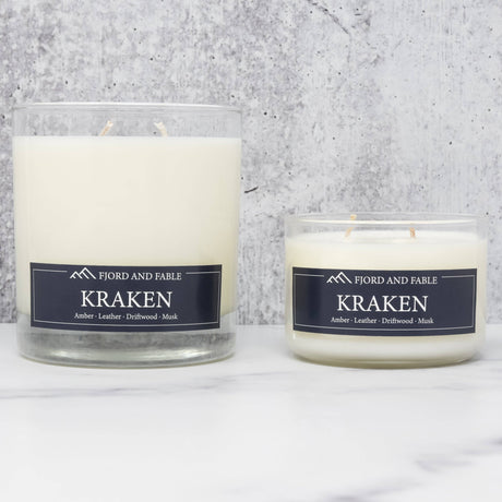 Two white soy wax candles in glass jars labeled “Fjord & Fable Candle: Kraken - Amber, Sea Salt, Driftwood & Leather 8oz,” set on white marble with a gray backdrop—perfect for adding bold amber and sea salt scent to any space.