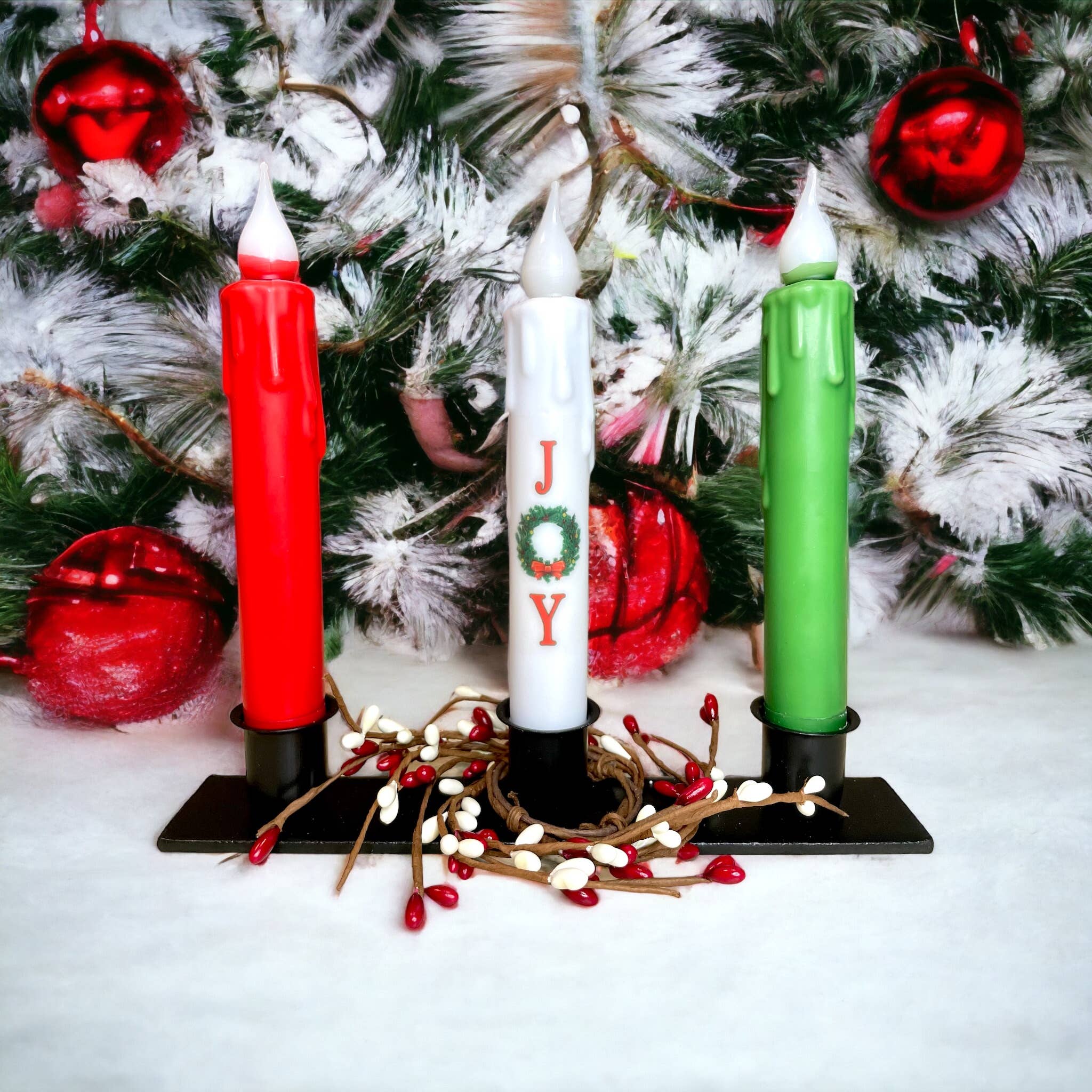 The Triple Wrought Iron Taper Holder beautifully displays three candles in red, white, and green, with the word JOY elegantly scripted on the white candle. Pine branches and ornaments add a festive charm to this exquisite centerpiece.