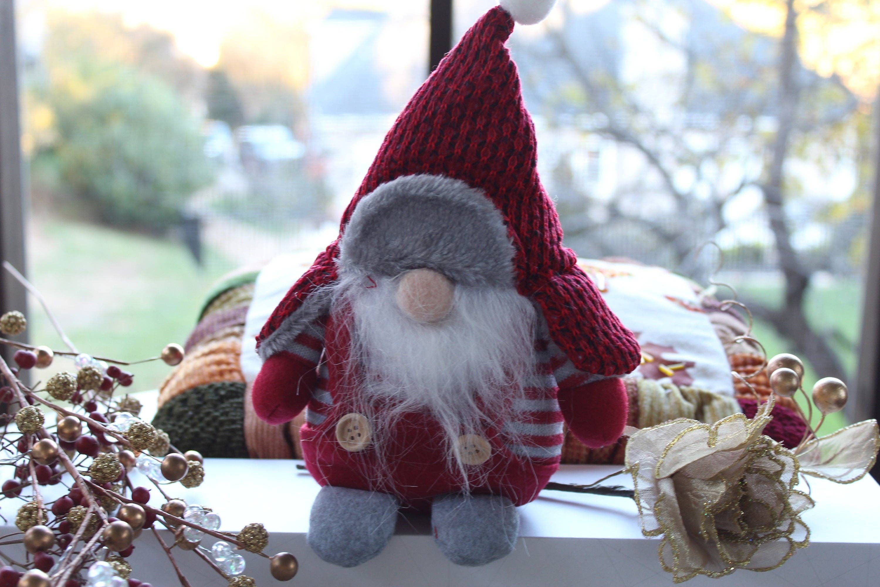 A Gnomes: Winter Gnomes Christmas plush toy, featuring Aldrich or Bernard with a long white beard, red hat, and striped outfit, is perched on a windowsill with a blurred outdoor background. Holiday decorations are situated nearby.