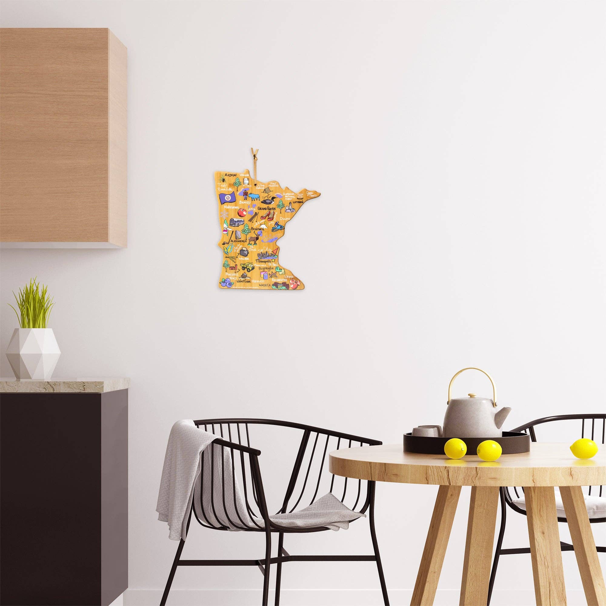 A modern kitchen boasts a Fish Kiss™ wooden wall decoration shaped like Minnesota, adorned with vibrant illustrations of landmarks. On the round table sit two bowls of yellow balls and a tea kettle, accompanied by the charming Minnesota Cutting Board featuring Fish Kiss™ artwork that adds an extra touch of warmth.