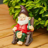 Garden gnome figurine with a red shirt and green pants sitting barefoot in a wooden chair, holding a green bottle, surrounded by leafy plants.