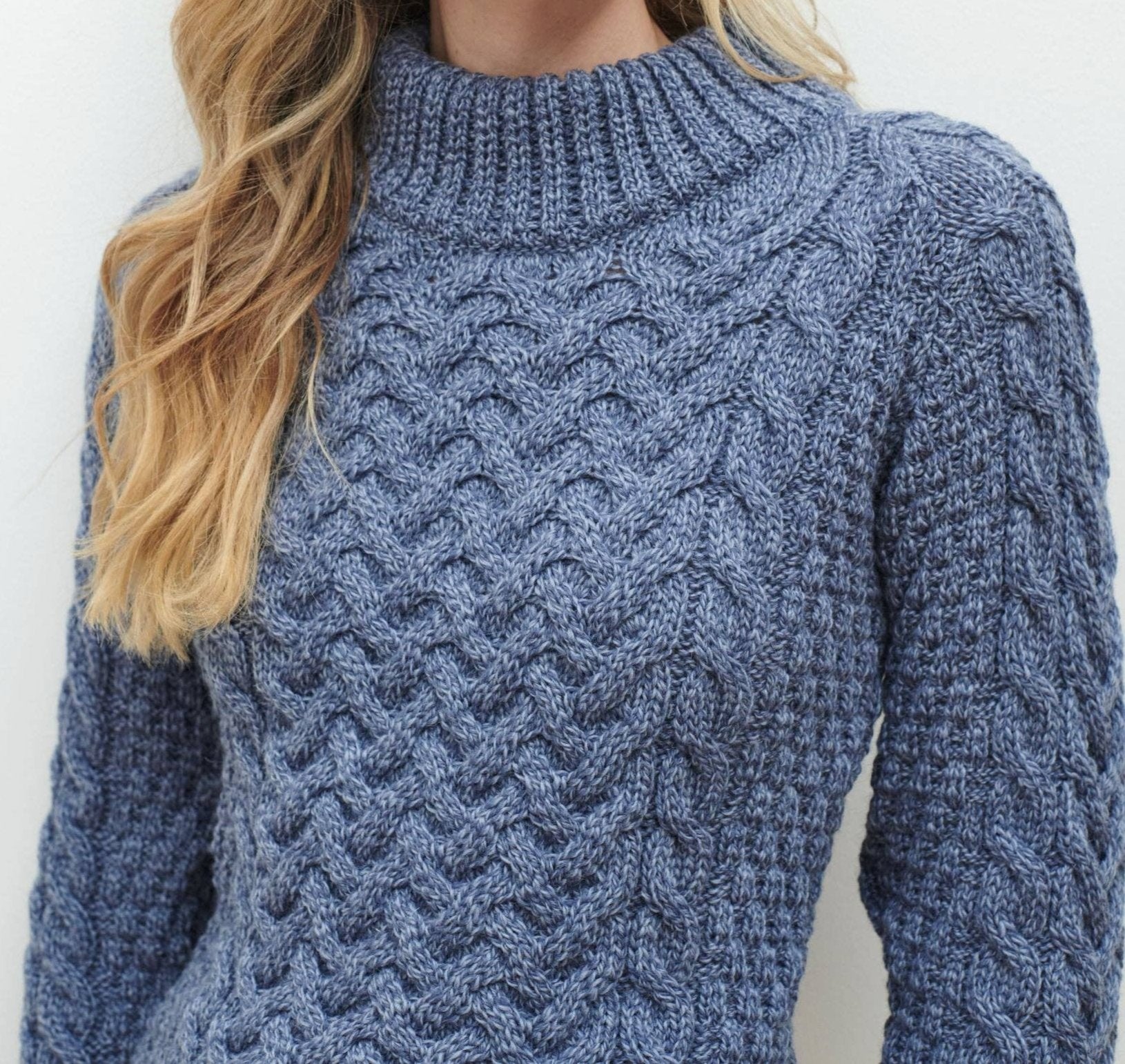 A person is wearing the Knightstown - Aran Crew Sweater in Denim, a textured cable stitch sweater made from soft Merino wool, featuring long sleeves and a high neckline.