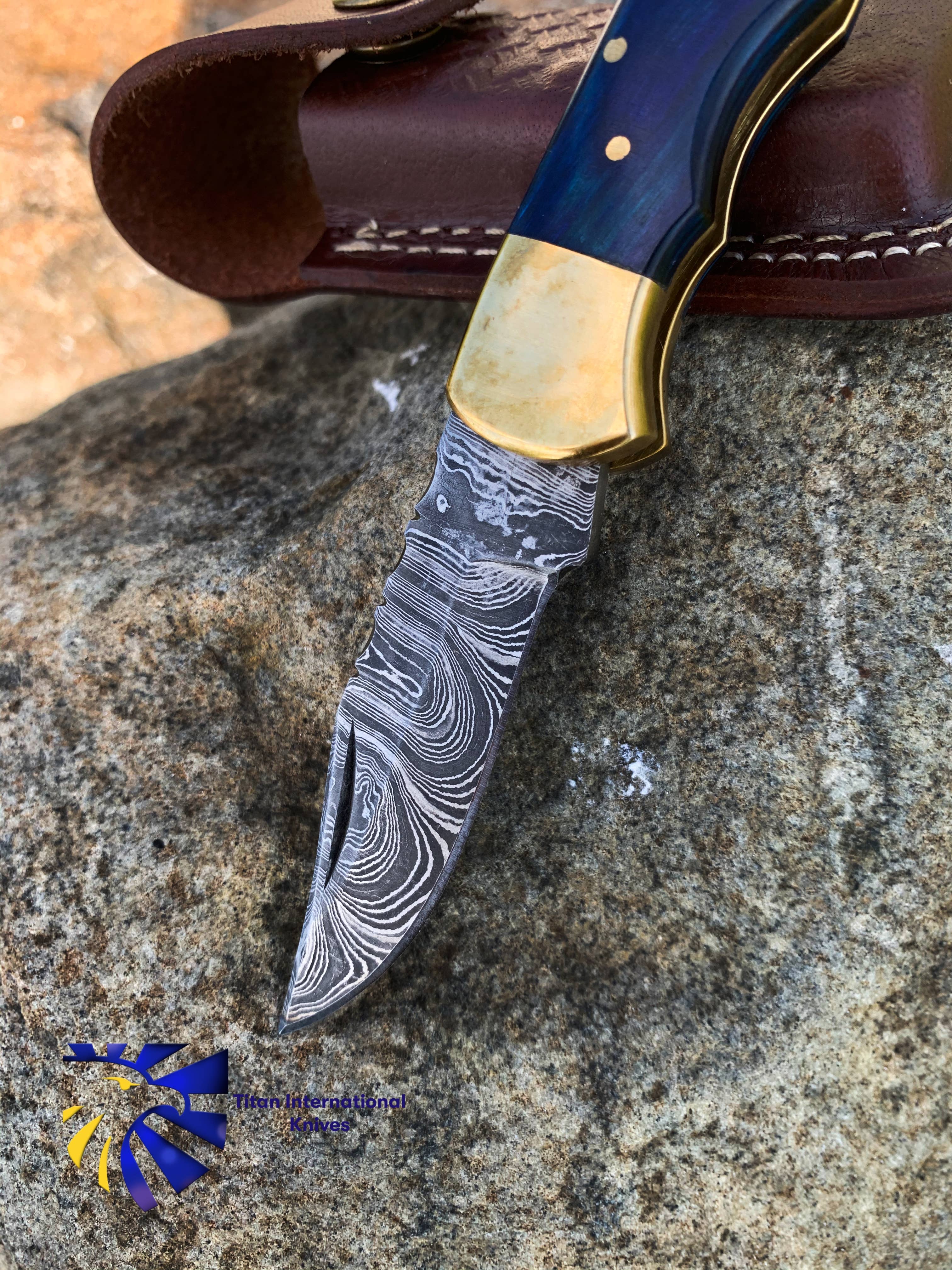 The Blue Diamond Wood Folding Knife w/ Leather Sheath is open on a rock, highlighting its Damascus steel blade, brass bolster, sturdy back lock, and intricate pattern, with the leather sheath resting nearby.