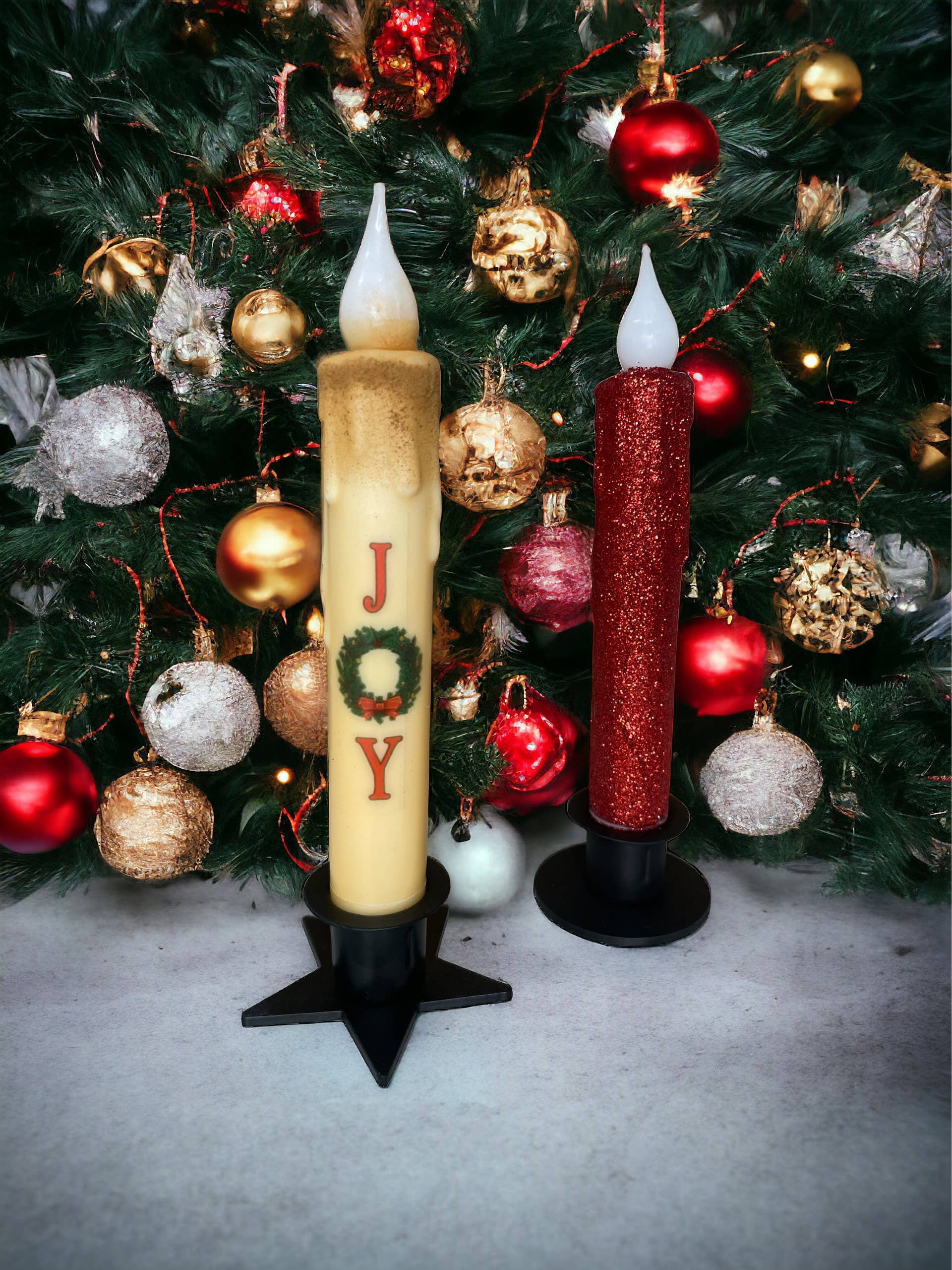 Two large decorative candles rest elegantly on Star Wrought Iron Taper Holders, placed in front of a Christmas tree adorned with red, gold, and silver ornaments. The yellow candle features JOY text, while the red one is glittery, adding a festive touch.