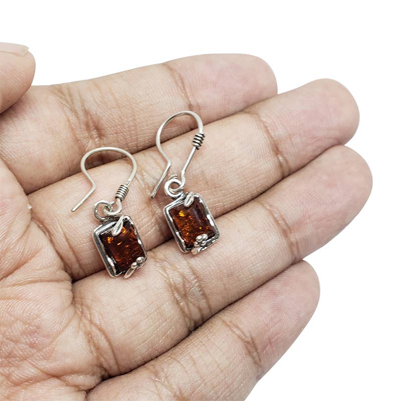 A hand cradles two .925 sterling silver earrings, each set with genuine Cognac Baltic amber stones in a captivating rectangular shape.