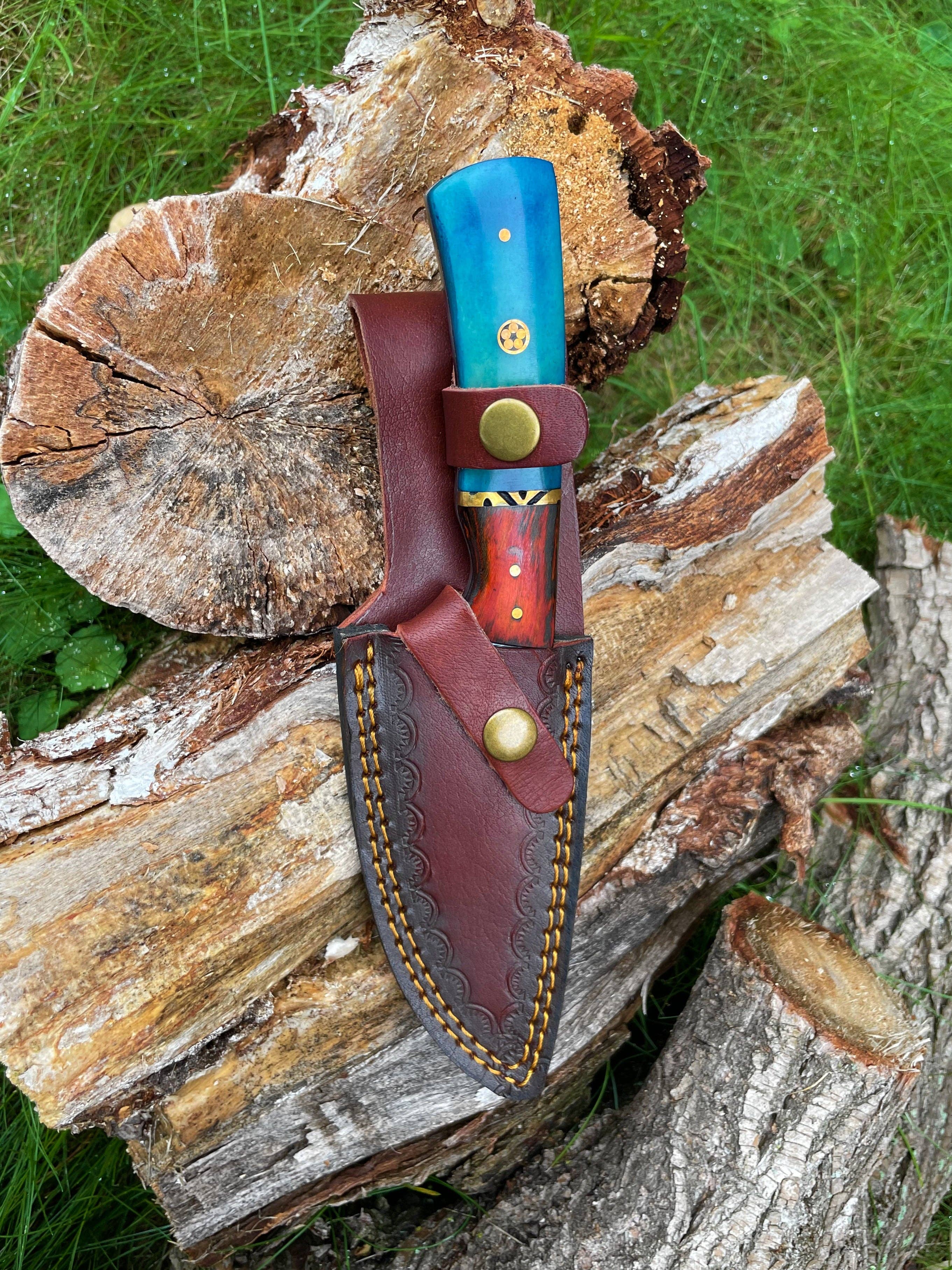 A hand forged Damascus knife with a black drop-style blade rests in a brown leather sheath on stacked wood outdoors.