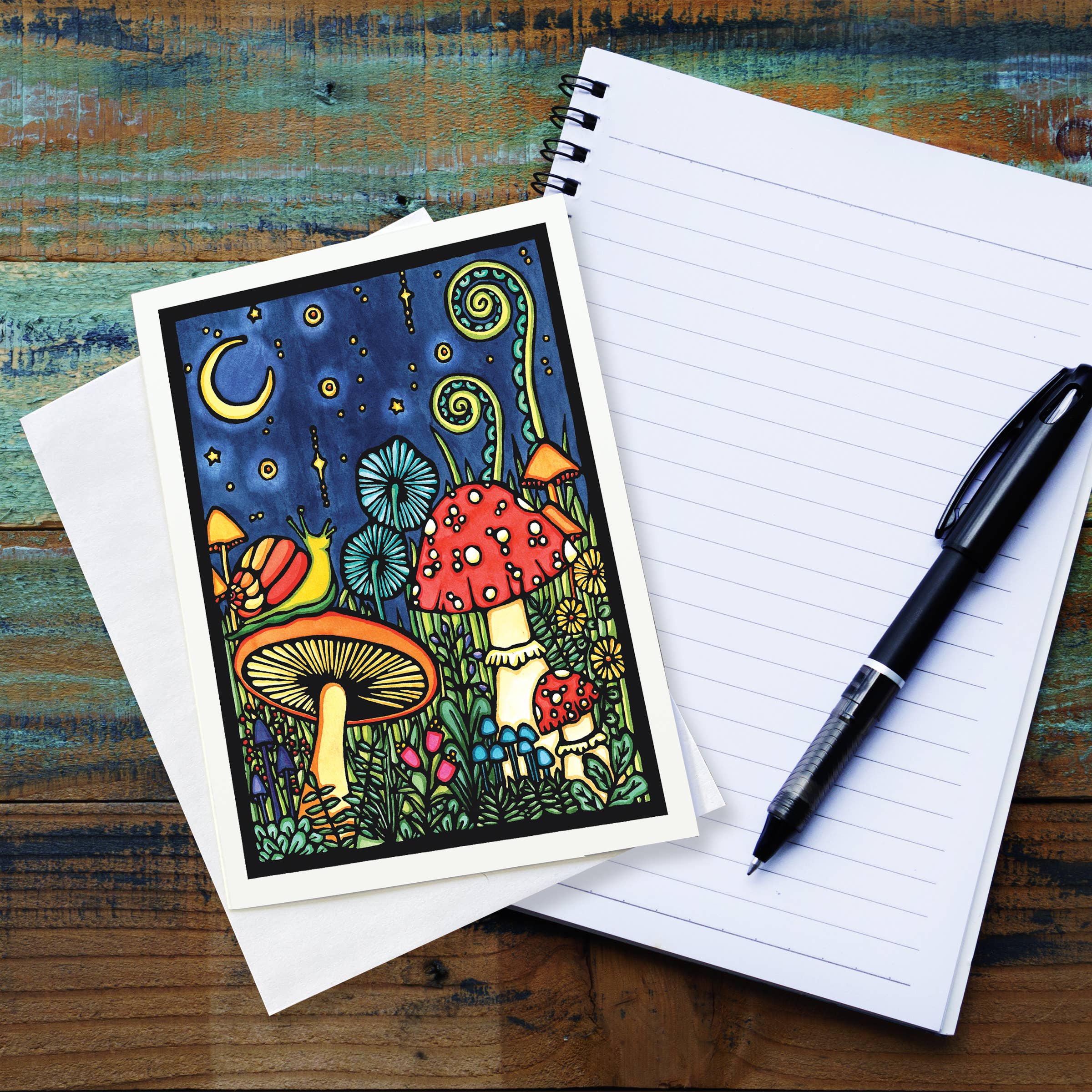 A Greeting Card: Mushroom Greeting Card with vibrant mushrooms and plants, styled after Sarah Angst Art, rests on a blank lined notebook. A black pen is ready to capture thoughts inspired by whimsical linocut prints.