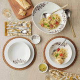 A table set for two with pasta, bread, wine, and utensils on leaf-designed plates, placed on Natural Grass Braided Woven Placemats and patterned napkins.