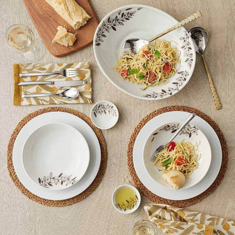A table set for two with pasta, bread, wine, and utensils on leaf-designed plates, placed on Natural Grass Braided Woven Placemats and patterned napkins.