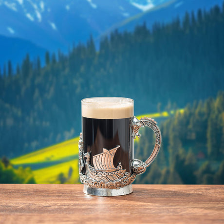 An 18 oz Mug: Viking & Mythological Pewter Glass with a Viking ship holder and pewter medieval engravings rests on wood, mountains and pine trees softly blurred in the background.