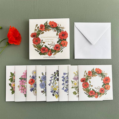 Koustrup & Cos Summer Wreaths card pack features 8 unique floral-themed cards with envelopes, beautifully displayed on a green surface next to a red poppy. The elegant packaging provides an enchanting square card folder for organization.