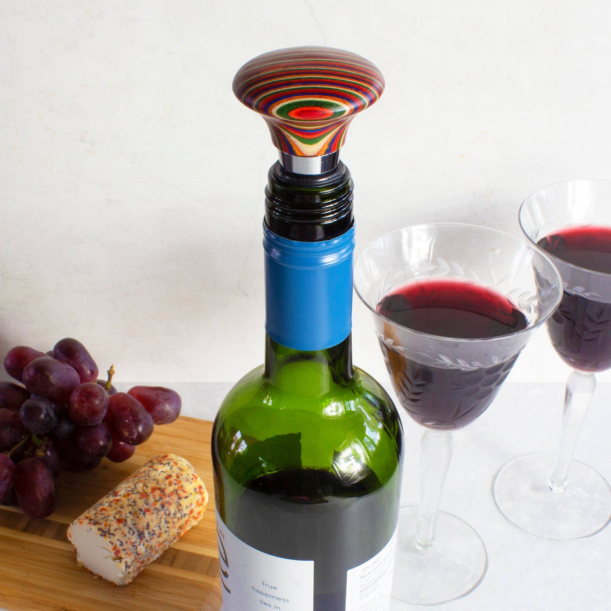 A wine bottle featuring the Marrakesh Collection Wine Bottle Stopper rests on a table, accompanied by two filled glasses, grapes, and a log of herbed cheese. The stopper includes air-tight silicone rings to preserve the fresh flavor.