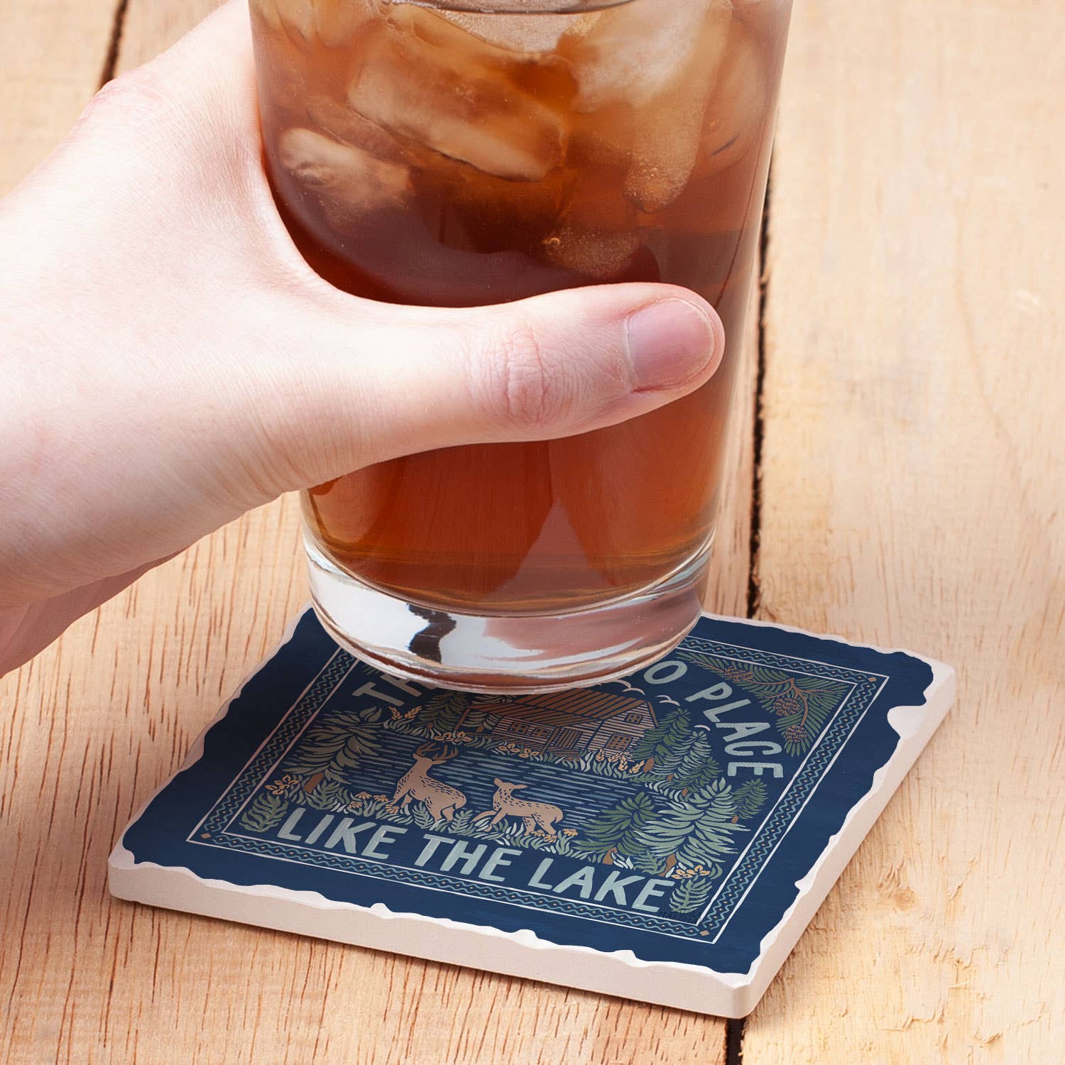 A hand holds a refreshing glass of iced tea on the Lake Place single tumbled tile coaster. This absorbent coaster, with decorative text and nature-themed illustrations, features a cork backing for stability on the wooden table.