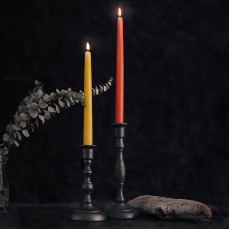 Two eco-friendly, dripless beeswax taper candles, one yellow and one red, are lit in dark holders on a table with a dried plant in a vase and driftwood against a dark backdrop, creating an elegant ambiance.