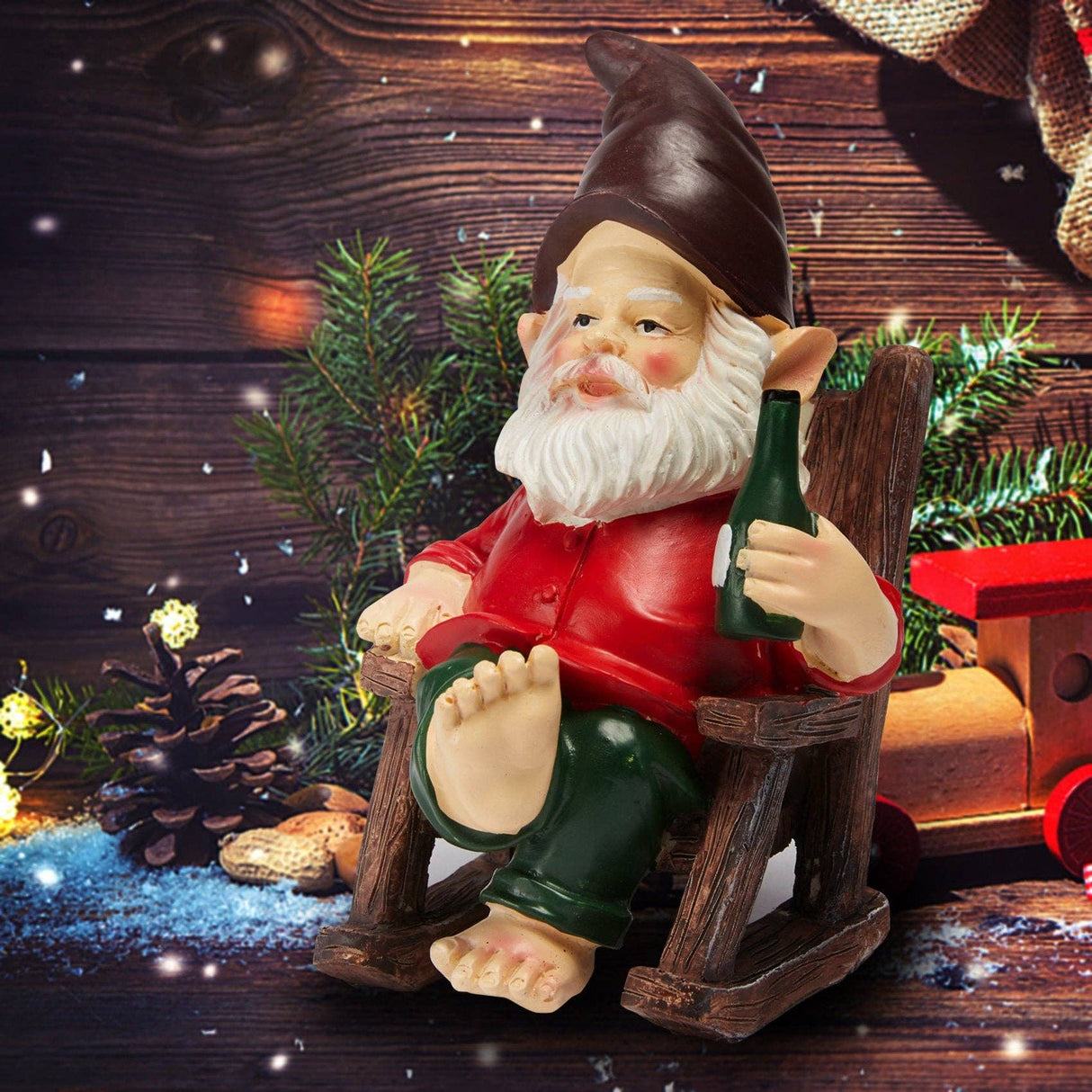 Gnome figurine in red shirt and green pants, barefoot on a wooden chair, holding a bottle. Pine branches, artificial snow, and a red toy train are in the background.