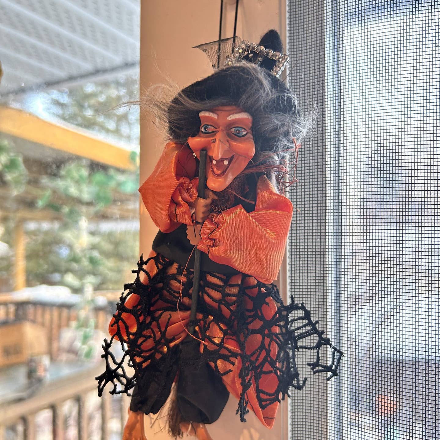 The Decor: Kitchen Witch Doll Sirene Orange 10.2" Tall, dressed in orange and black with a pointed hat, hangs by a window as a good luck charm, with trees and a building seen outside.