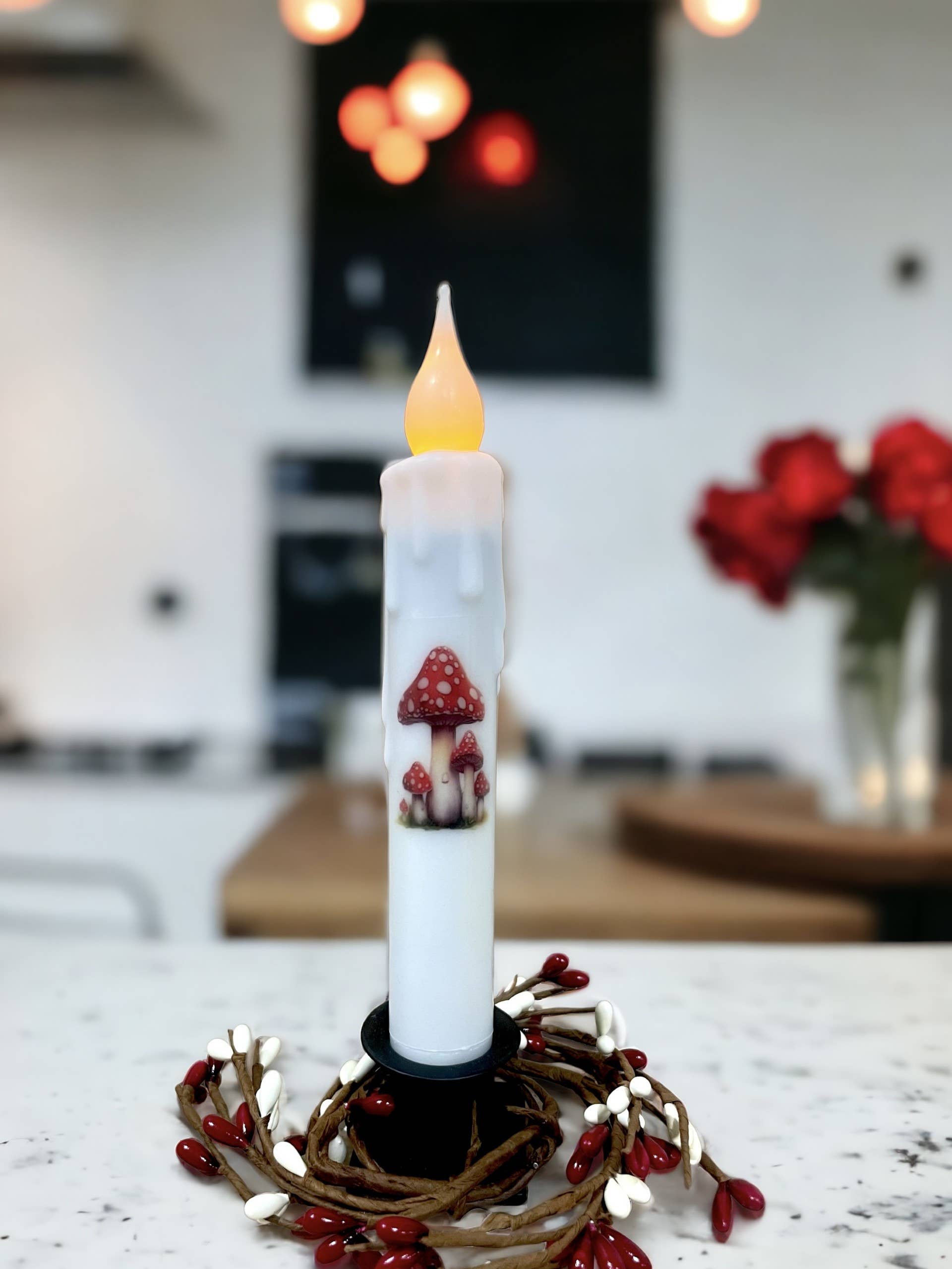 The Candle: Red Spotted Mushroom Battery Operated Timer Taper Candles are tall white LED candles with flame-like bulbs, showcasing red spotted mushroom designs on a decorated base with red and white accents, against a blurred home interior background.