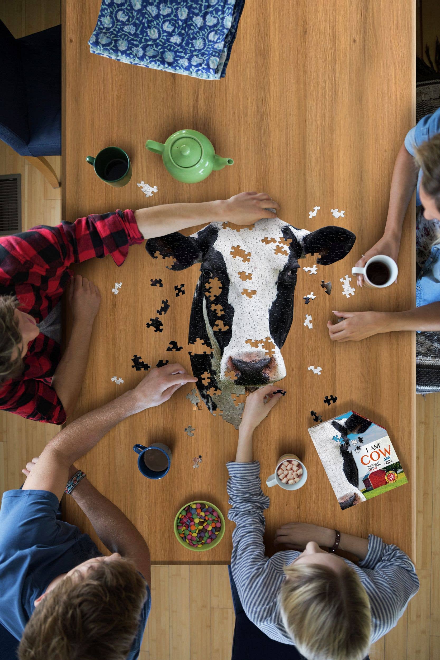 Four people are absorbed in putting together the I AM Cow 300-piece puzzle, crafted from premium quality chipboard, on a wooden table. Nearby, a green teapot, cups, and an educational Fun Facts Cow book enhance the ambiance, creating an enriching and enjoyable experience for everyone involved.