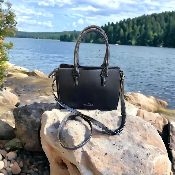 A Harris Tweed Double Handle Tote in grey is placed on a large rock by a lakeside, with trees and blue sky in the background.