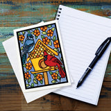 The Card: Birdhouse 5x7 Greeting Card by Sarah Angst features two birds and a birdhouse, comes with an envelope, is blank inside, and is made in America. Shown beside a spiral notebook and pen on a wooden table.