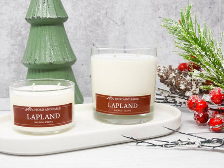 Two white Lapland Candle 8 oz glass jars by Fjord & Fable sit on a white tray beside a green tree ornament and festive greenery, filling the air with a cozy, woodsy balsam fir, clove, and cedar scent.