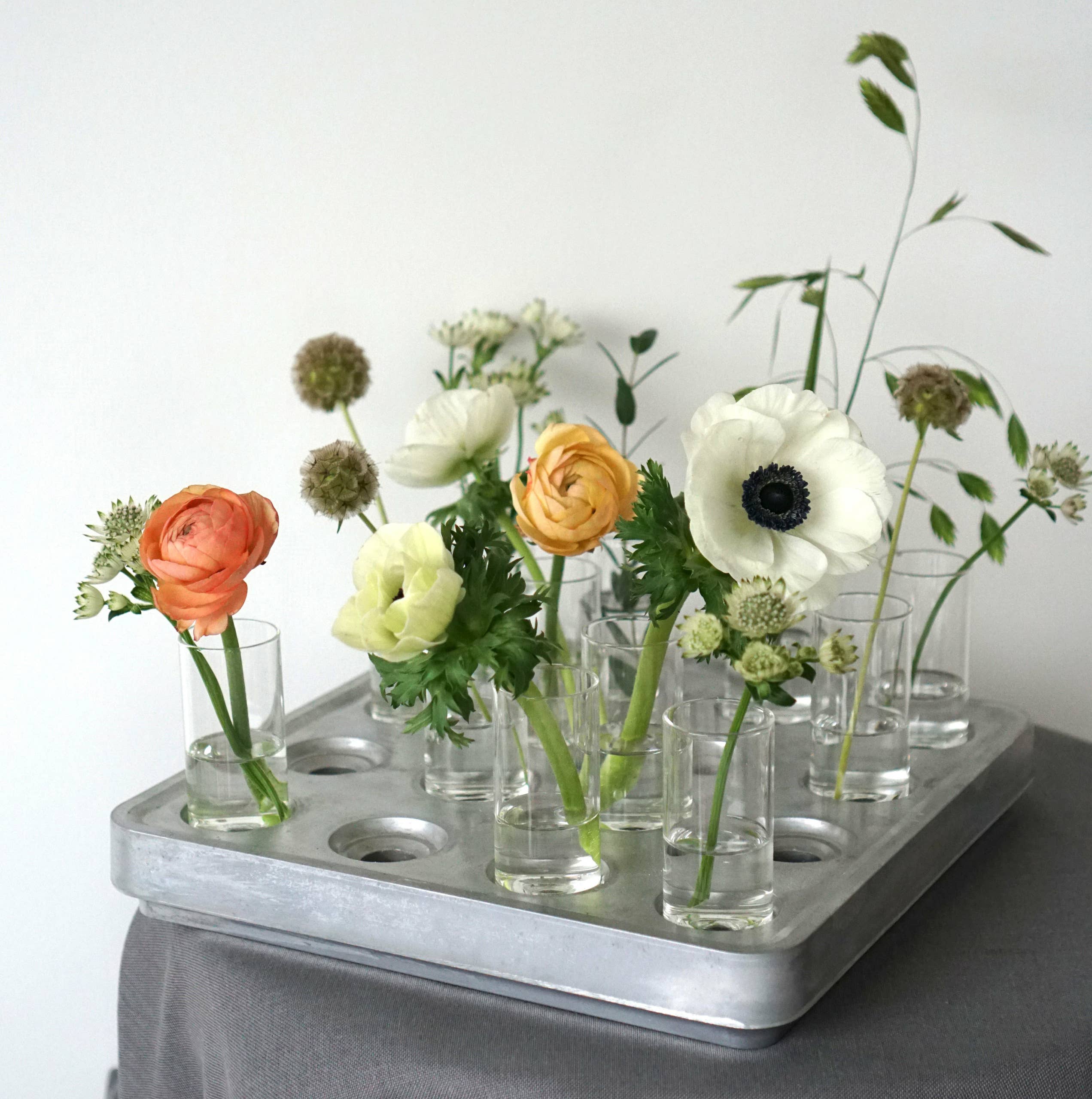 Mini vases filled with flowers sit on a metal tray alongside a Candle Holder: Mini Vases Pack of 4 for Stumpastaken, elegantly arranged on a gray cloth.
.