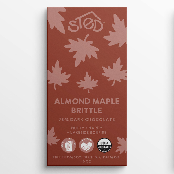 Rust-colored chocolate bar wrapper with pink maple leaf pattern, text indicating almond maple brittle and 70% dark chocolate, organic and allergy-friendly icons visible.