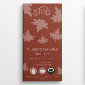 Rust-colored chocolate bar wrapper with pink maple leaf pattern, text indicating almond maple brittle and 70% dark chocolate, organic and allergy-friendly icons visible.